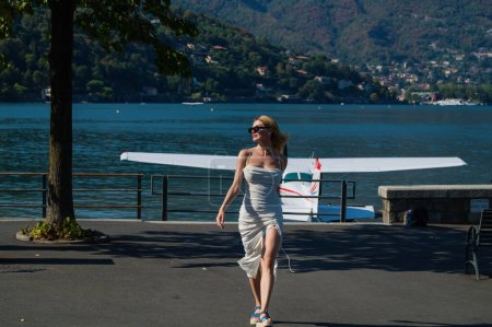 Téléchargez les photos : Femme marchant sur le lac de Côme, Italie. Fille dans une robe sexy près du lac de Côme. Vacances d'été italiennes. Femme de la mode dans la rue près de jet plane. Jeune voyageuse en vacances d'été à Côme - en image libre de droit