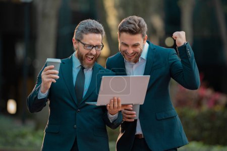 Photo for Excited business men team using laptop outdoor. Businessmen looking laptop with their business success in city background. Freelance concept. Two men talking in urban city center working on laptop - Royalty Free Image