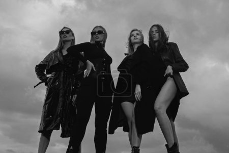 Téléchargez les photos : Portrait de mode de filles de modèles de mode de groupe posant en plein air, tenue de style noir contre le ciel. Jeunes femmes attrayantes en vogue - en image libre de droit