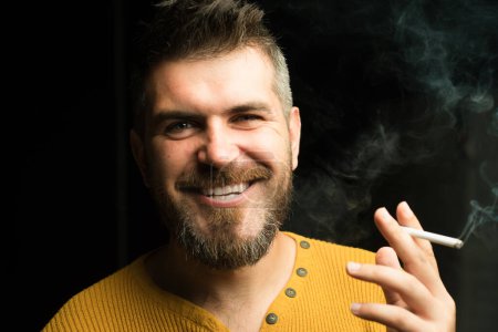 Foto de Smiling man smoking cigarette. Smoke on black background. Smoking addiction. Cigarette nicotine addiction. Smoke on black - Imagen libre de derechos