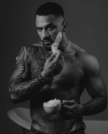 Téléchargez les photos : Bearded handsome tattooed man apply a shaving foam at his face. Bath procedures and grooming. Brutal muscular man going to shave his beard. Man style and fashion concept - en image libre de droit