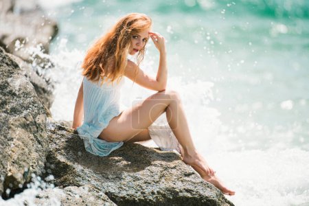 Photo for Summer dream. Sexy woman sitting on rocky beach against sea blue waves. Sensual young woman wearing sexy summer dress on a beach with her elbow on her knee and her hand to her head - Royalty Free Image