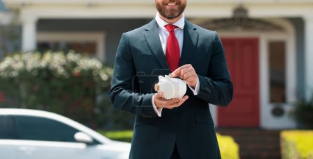 Photo for Happy young man putting money in piggy bank. Businessman holding piggybank on rich house background. Save money and financial investment - Royalty Free Image