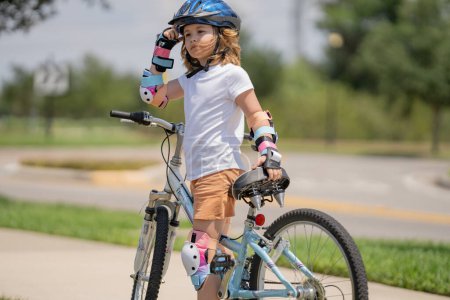 Foto de Un niño pequeño montando en bicicleta en el parque de verano. Los niños aprenden a conducir una bicicleta en un camino de entrada afuera. Niño montar en bicicleta en la ciudad con cascos como equipo de protección. Niño en bicicleta al aire libre - Imagen libre de derechos