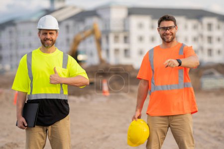 Téléchargez les photos : Deux travailleurs sur le chantier de construction travaillant sur un casque à l'extérieur. Travailleurs des constructeurs travaillant sur le chantier. Le travail d'un travailleur sur le chantier exige beaucoup de force physique - en image libre de droit
