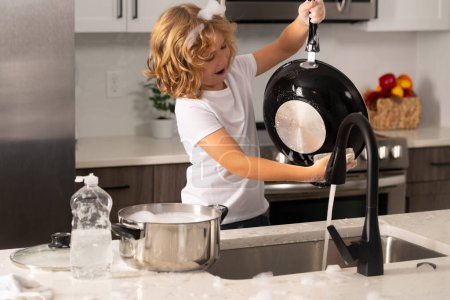 Foto de Niño lavar los platos en el interior de la cocina. Niño ayudando a sus padres con las tareas domésticas. Un niño lindo lavando platos cerca del fregadero en la cocina - Imagen libre de derechos