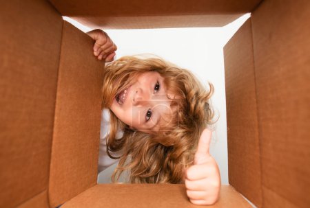 Photo for Child opens carton box and pulling out gift from it. Small boy looking in parcel box and happy to receiving a surprise. Young kid showing joy on his face. Low angle view - Royalty Free Image
