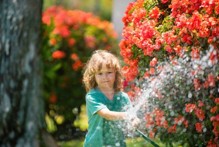 Téléchargez les photos : Arrosage des fleurs et des plantes dans le jardin. Enfant avec tuyau d'eau dans la cour. Jardinage des enfants. Enfants plaisir d'été en plein air à la maison - en image libre de droit