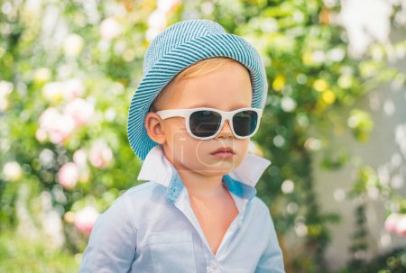 Téléchargez les photos : Mode pour enfants. Joyeux enfant en lunettes de soleil et chapeau, adorable adorable enfant. Joyeux portrait de petit enfant sur fond vert nature à l'extérieur - en image libre de droit