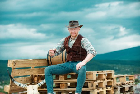 Téléchargez les photos : Un paysan cowboy boit un whisky à la campagne avec un chapeau de cow-boy occidental. Américain Homme modèle dans la campagne à la ferme - en image libre de droit