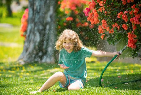 Téléchargez les photos : Mignon petit garçon arrosant des fleurs dans le jardin le jour de l'été. Enfant agriculteur avec tuyau d'arrosage dans la plantation de plantes. tuyau. drôle enfant arrosage des plantes dans le jardin de la cour - en image libre de droit