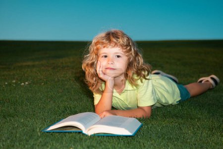 Téléchargez les photos : Enfant garçon lecture livre, étendu sur l'herbe dans le champ sur fond de ciel. Portrait d'enfants intelligents - en image libre de droit
