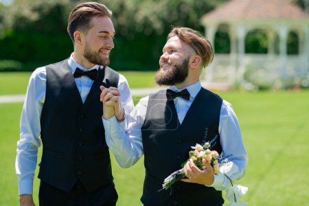Photo for Gay grooms walking together on Wedding day. Married LGBT couples celebrate a romantic wedding ceremony together with a bouquet flower. Portrait of happy gay couple on their wedding day outdoor - Royalty Free Image