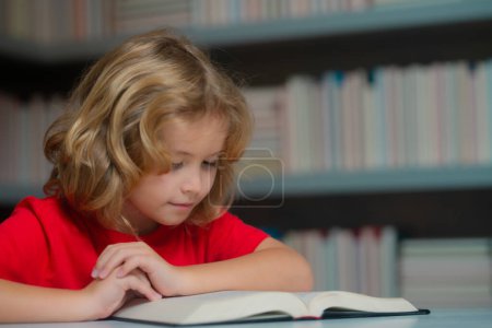 Photo for Little student boy reading book at school. Kid doing homework, sitting at table by books, in library - Royalty Free Image