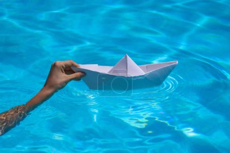 Foto de Mano femenina sosteniendo barco de papel sobre el fondo del mar. Origami barco de papel navegando en el agua causando olas y ondulaciones. Concepto de turismo, viajes sueños vacaciones vacaciones - Imagen libre de derechos