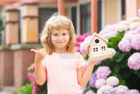 Foto de Lindo niño jugando con el modelo de casa pequeña al aire libre en el jardín en casa. Casa de ecología en manos de niños contra patio trasero - Imagen libre de derechos
