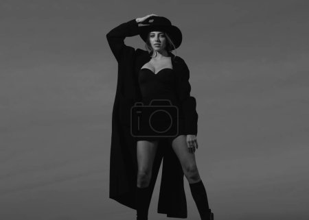 Téléchargez les photos : Femme de mode en manteau de mode noir, chapeau et chaussures noires bottes. Femme de mode posant à l'extérieur - en image libre de droit