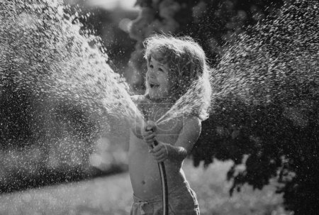 Téléchargez les photos : Enfant jouant dans le jardin, verse du tuyau, fait une pluie. Concept d'enfance heureuse - en image libre de droit