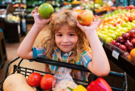 Foto de Niño eligiendo una manzana en una tienda. Gracioso niño sonríe y sostiene la manzana. Un chico de compras en el supermercado. Pequeño niño eligiendo verduras frescas en una tienda de alimentos - Imagen libre de derechos