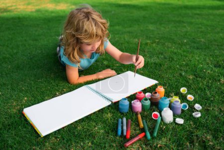 Téléchargez les photos : Les enfants créent artiste peint vacances créativité. Kid dessine dans le parc posé dans l'herbe en s'amusant sur fond de nature - en image libre de droit