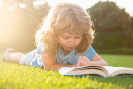 Foto de Kid boy reading a book lying on grass. Cute little child in casual clothes reading a book and smiling while lying on grass in park. Smart clever Kids - Imagen libre de derechos