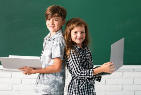 Foto de Niños de primaria. Retrato de niños niño y niña con computadora portátil y libro en la escuela. Niños en clase en pizarra - Imagen libre de derechos