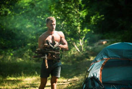 Foto de Fuerte hombre sexy con leña para hoguera. Camping al aire libre en la naturaleza. Hombre leñador guapo y musculoso con un hacha, acampando. Caminata de fin de semana - Imagen libre de derechos