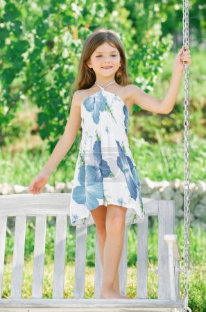 Téléchargez les photos : Mode enfants robe. Bonne petite fille sur une balançoire dans le parc d'été. Enfant heureux s'amuser à l'extérieur - en image libre de droit