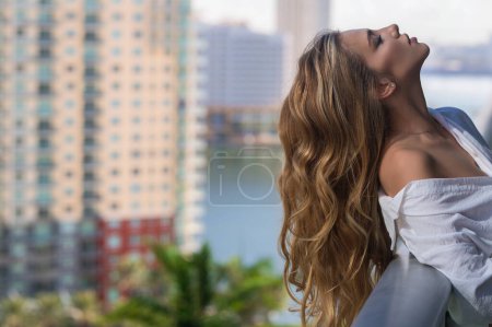Photo for Woman relaxing on balcony. Beauty woman in stylish sexy shirt on balcony enjoying view on big city in Miami. Leisure morning time and lifestyles concept. Relaxing girl on balcony and extends long hair - Royalty Free Image