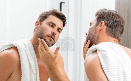 Foto de Hombre hispano de mediana edad mirando en el espejo, la piel facial y rastrojos. Producto masculino del cuidado de la belleza. Cuidado de la piel, spa en casa. Retrato de belleza de un hombre hermoso. Modelo de spa, crema hidratante nutritiva - Imagen libre de derechos