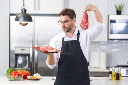 Téléchargez les photos : Chef homme cuisine viande steak de bœuf dans la cuisine. Portrait d'un homme occasionnel cuisinant avec des ingrédients de viande. Homme décontracté préparant de la viande crue dans la cuisine. Chef cuisinier steak de boeuf - en image libre de droit