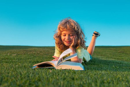 Foto de Chico gracioso leyendo el libro en el parque de primavera. Niño leyendo libro, tendido en la hierba en el campo en el fondo del cielo. Retrato de niños inteligentes - Imagen libre de derechos