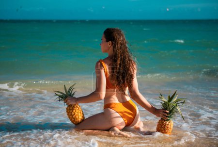 Téléchargez les photos : Female buttocks in thongs bikini, sexy ass. Young woman holding a pineapple on sea sand beach background - en image libre de droit