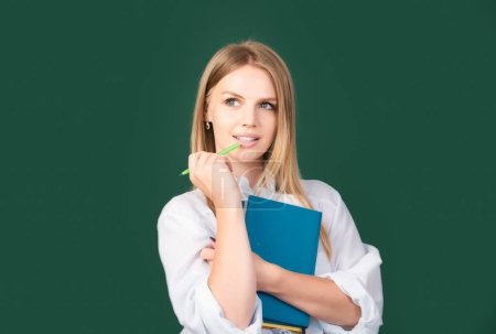 Photo for Portrait of young cute blonde female college student studying in classroom on class with blackboard background. Female student taking notes from a book in college. Thinking young woman - Royalty Free Image