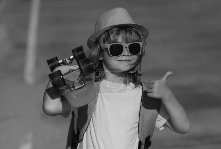 Téléchargez les photos : Enfant étonnant avec pouce levé, portrait de rue. Petit garçon 7-8 ans tenir skateboard posant sur la rue en plein air. Concept de style de vie de l'enfance. Skateboard pour enfants - en image libre de droit