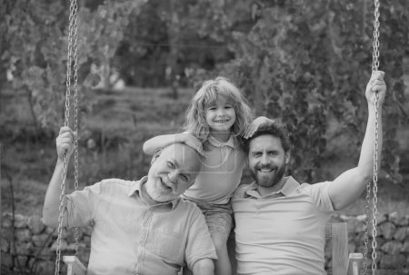 Téléchargez les photos : Trois générations d'hommes ensemble, portrait d'un fils, d'un père et d'un grand-père souriants tournant sur la balançoire, s'amusant dans le parc en plein air - en image libre de droit