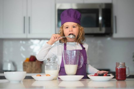 Téléchargez les photos : Chef enfant lécher cuillère cuisine repas sain et préparation du dîner. Chef cuisinier enfant apprend à faire un gâteau dans la cuisine de la maison - en image libre de droit