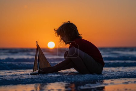 Téléchargez les photos : Concept d'aventure et de voyage. Enfant jouant avec bateau à voile jouet sur fond de mer d'été. Voyages et vacances d'été. Enfant jouant avec le bateau jouet dans l'eau sur la plage. Activités d'été en plein air - en image libre de droit