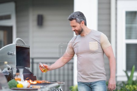 Téléchargez les photos : Concept de barbecue. Homme hispanique d'âge moyen en t-shirt pour barbecue. Torréfaction et grillage des aliments. Faire rôtir la viande à l'extérieur. Barbecue et grill. Cuisson de viande dans la cour arrière - en image libre de droit