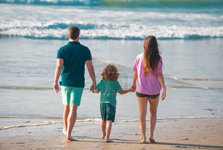 Foto de Vista trasera de la feliz familia joven caminando en la playa. Niño con padres tomados de la mano. Poeple de cuerpo entero. Viajes en familia, concepto de vacaciones - Imagen libre de derechos