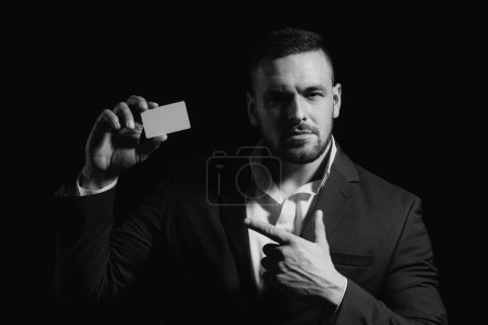 Téléchargez les photos : Portrait d'un jeune homme vêtu d'un costume pointant du doigt une carte de crédit isolée sur fond noir. Homme d'affaires avec carte de crédit - en image libre de droit