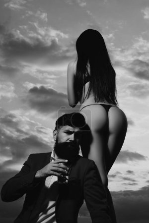 Photo for Bearded businessman in elegant suit with glass of whiskey. Rich man drink. Sencual couple in love. Womans butt in bikini - Royalty Free Image