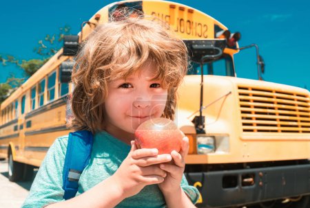 Foto de El chico de la escuela en el frente del autobús escolar comiendo manzana. Niño de la escuela primaria con bolsa en el autobús escolar backgroung - Imagen libre de derechos