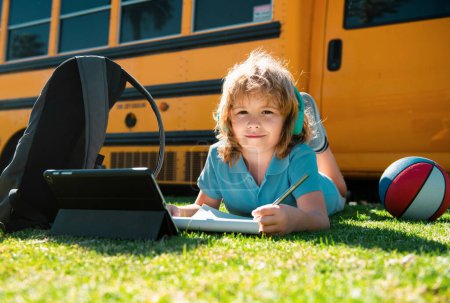 Foto de Alumno escribiendo tarea en la hierba con la tableta portátil al aire libre cerca del autobús escolar. Aprender en línea, concepto de educación infantil - Imagen libre de derechos