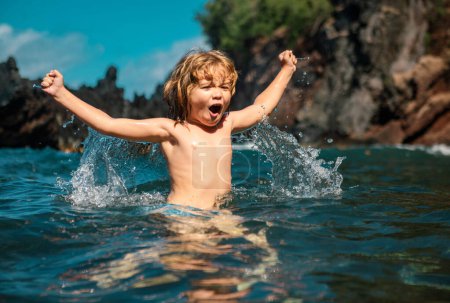 Foto de Niño sorprendido jugando y salpicando en el mar. El chico se divierte al aire libre. Vacaciones de verano y concepto de estilo de vida familiar saludable. Niño levanta las manos en el agua y salpica gotas de agua - Imagen libre de derechos