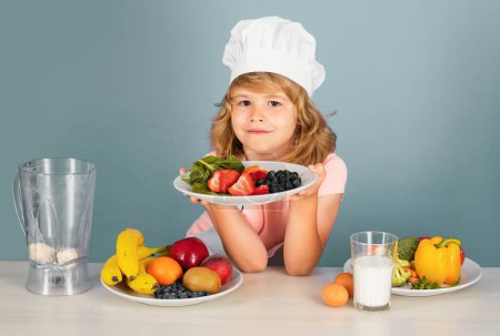 Téléchargez les photos : Assiette enfant chef avec fruits isolés sur bleu. Drôle petit chef cuisinier enfant portant casquette de cuisinier uniforme et tablier cuisiné dans la cuisine - en image libre de droit