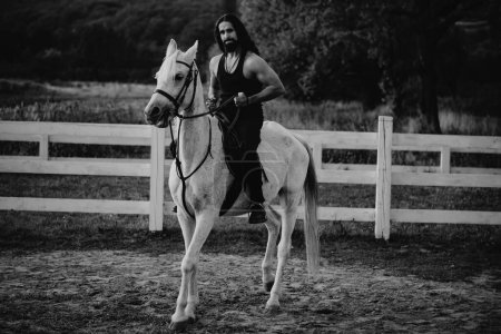 Téléchargez les photos : Homme attrayant assis sur un cheval blanc sur le ranch en automne. Pleine longueur de jeune homme beau assis sur son étalon à la campagne. Homme équestre sur son équitation à la nature - en image libre de droit