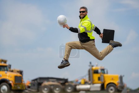 Photo for Crazy builder excited jump on site construction. Excited builder construction worker in a safety helmet jumping in front of the trucks. Excited crazy builder man in helmet jump outdoor - Royalty Free Image