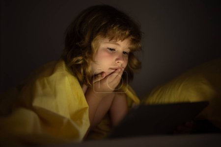 Foto de Niño acostado en la cama por la noche y jugando tableta digital. Niño viendo la tableta. Niño en la cama por la noche viendo la tableta, cara iluminada por la luz de la pantalla. Niño con la tableta en la cama viendo dibujos animados - Imagen libre de derechos