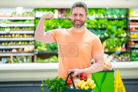 Photo for Handsome man hold shopping cart full of groceries. Man holds shopping basket. Store, shopping, sales and discounts. Male shopper. Man holding shopping basket with organic vegetables and fruits - Royalty Free Image